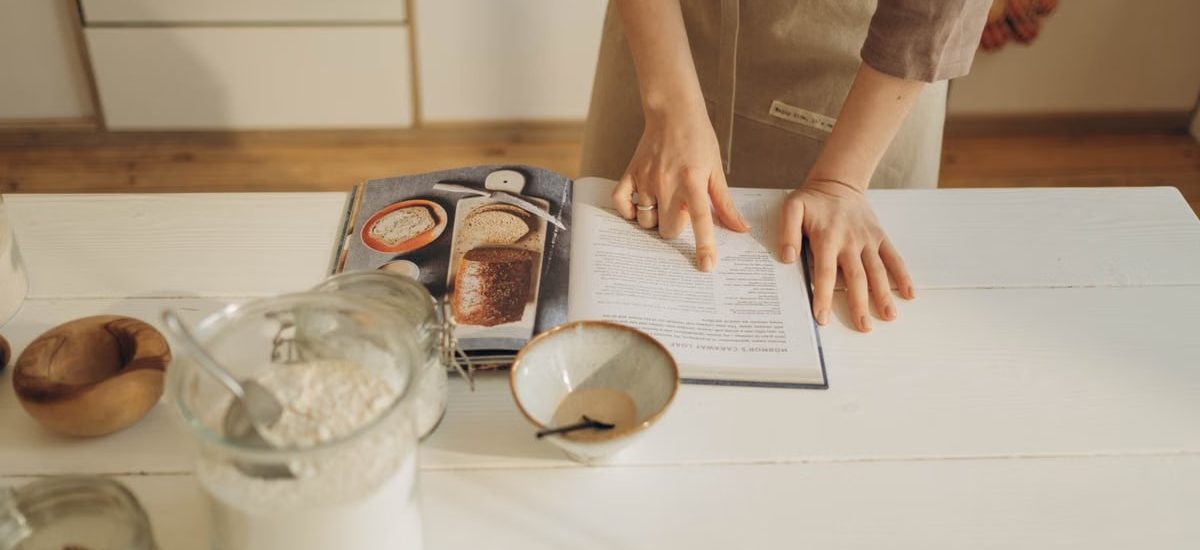 Hands pointing at cookbook in a cozy kitchen setting with ingredients ready for baking.