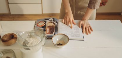 Hands pointing at cookbook in a cozy kitchen setting with ingredients ready for baking.
