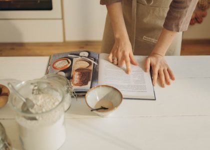 Hands pointing at cookbook in a cozy kitchen setting with ingredients ready for baking.